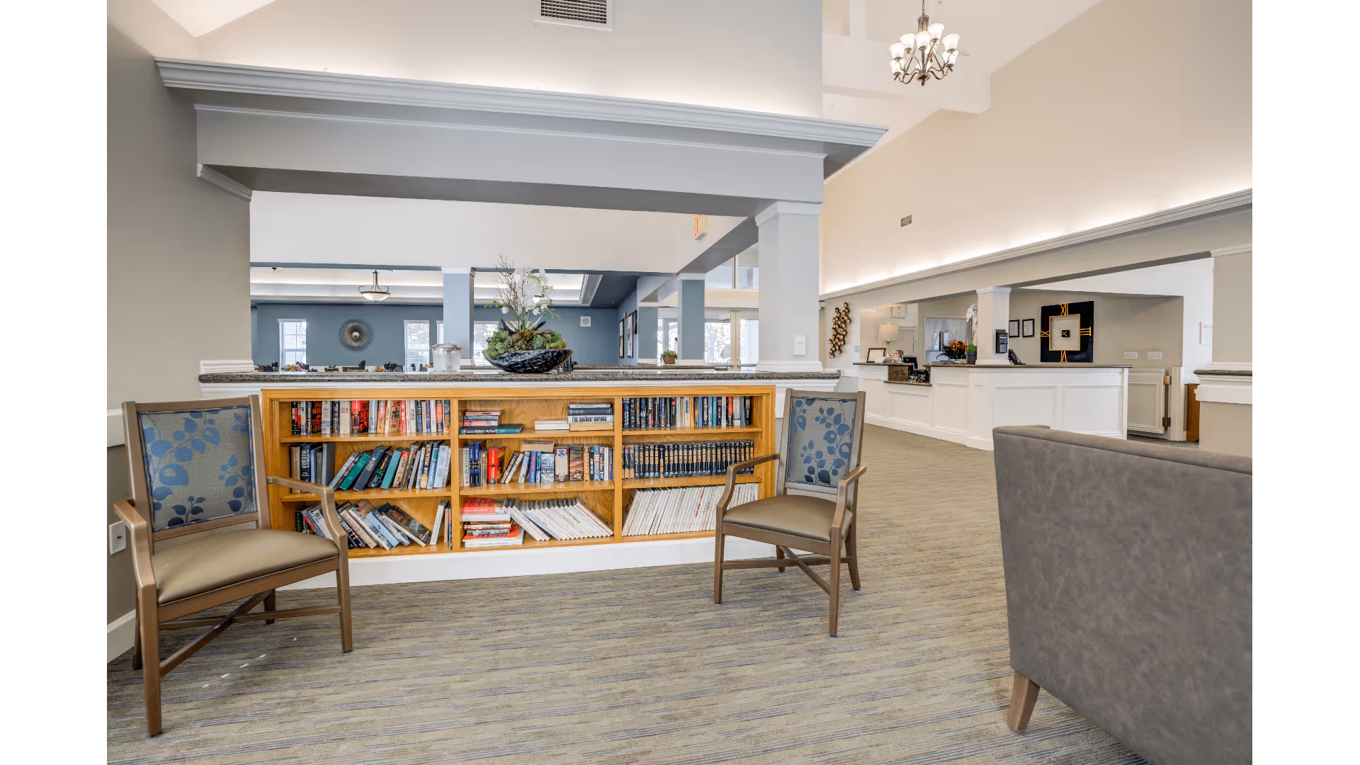 Interior view of a senior living facility lobby area with a built-in wooden bookshelf filled with books, two wooden chairs with patterned cushions on either side, and a gray sofa partially visible. The space has carpeted flooring, white walls with gray accents, and a chandelier hanging from the ceiling. In the background, there is a reception desk and additional seating areas.