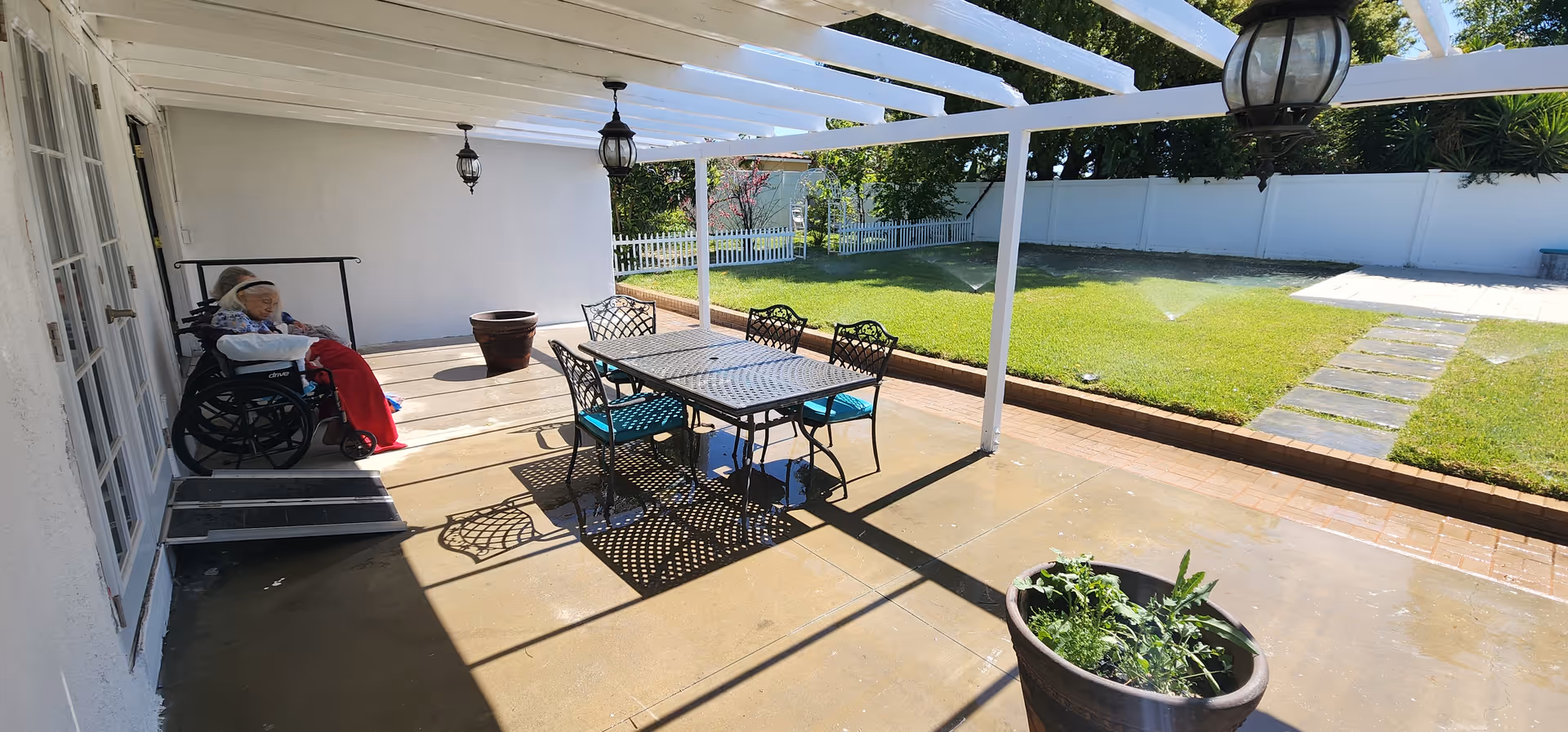 Outdoor patio area with a white pergola overhead, a metal table with four chairs, and two elderly individuals sitting near the wall, one in a wheelchair covered with a red blanket. The patio overlooks a green lawn with a sprinkler system and a white fence in the background.
