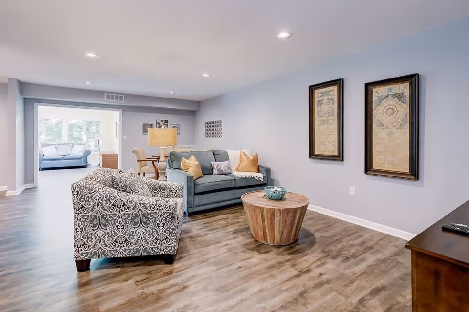A spacious living room area with light gray walls and wood flooring. The room features a patterned armchair, a blue sofa with beige and striped pillows, a round wooden coffee table with a decorative bowl, and two framed artworks on the wall. In the background, there is a dining area with a table and chairs, and another seating area with a blue couch near large windows letting in natural light.