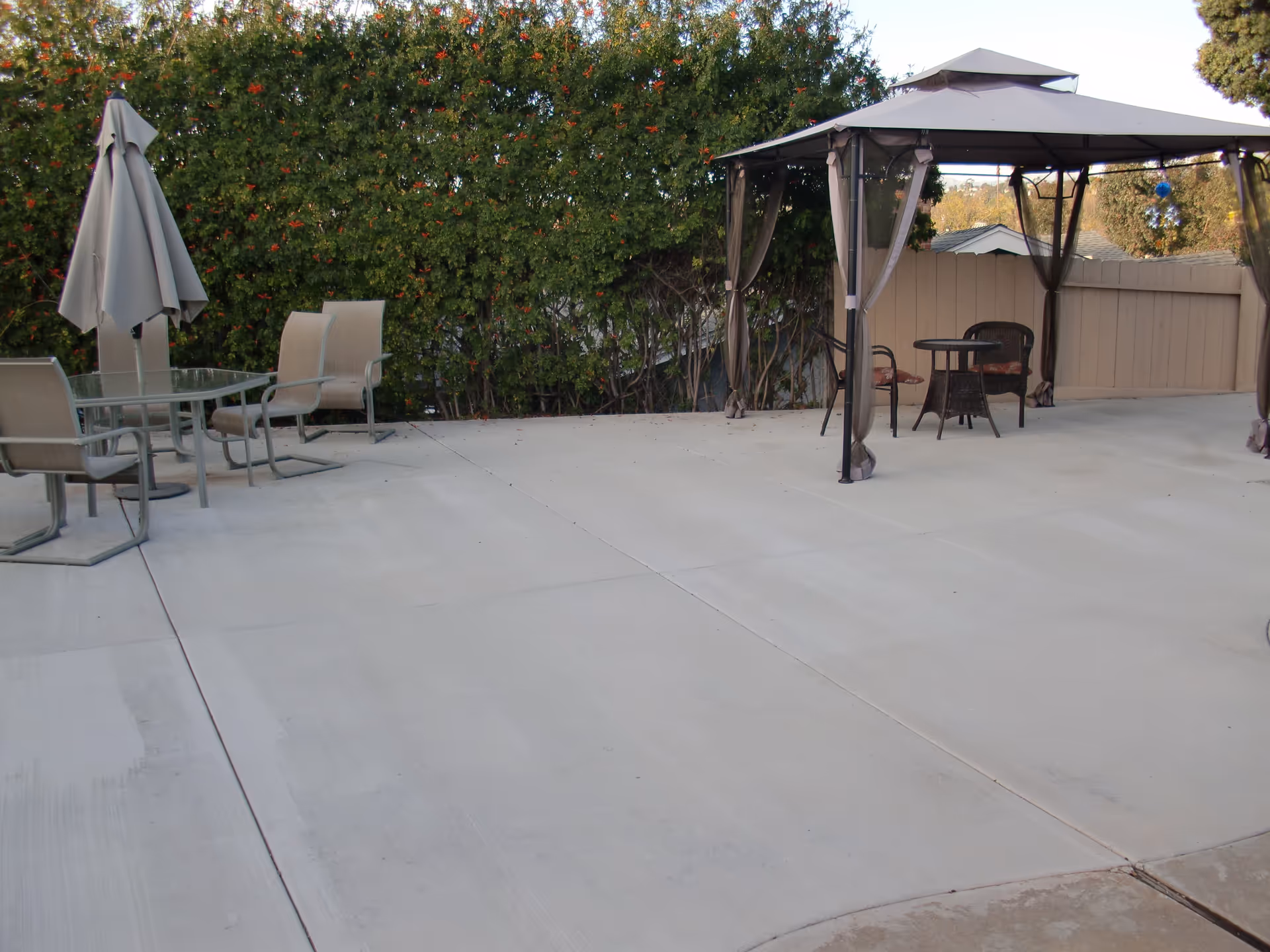 Outdoor patio area with concrete flooring, featuring a glass table with four beige chairs and a closed umbrella on the left side. On the right side, there is a gazebo with sheer curtains and a small round table with two chairs underneath. Green bushes and a wooden fence are in the background.