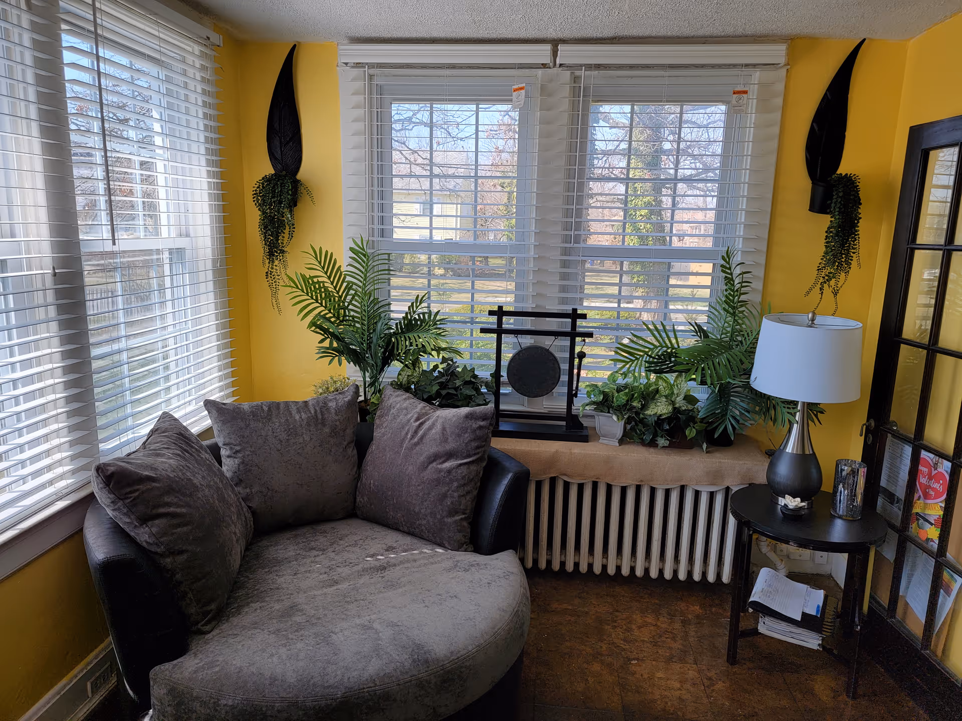 A cozy corner of a living room with a large round gray cushioned chair with two matching pillows. The walls are painted yellow and there are large windows with white blinds letting in natural light. Several green plants are placed on the windowsill and hanging on the walls. A small black side table with a lamp and some papers is next to the chair. A decorative gong is also placed on the windowsill.