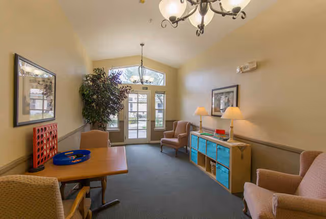 A cozy common area with beige walls and carpeted floor featuring a wooden table with chairs, a large plant near glass double doors, two armchairs, a wooden storage unit with blue bins, two table lamps, and framed artwork on the walls.