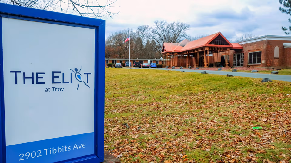 A freestanding sign reading "The Eliot at Troy" on 2902 Tibbits Ave stands on a lawn in front of the facility's brick building and entrance.