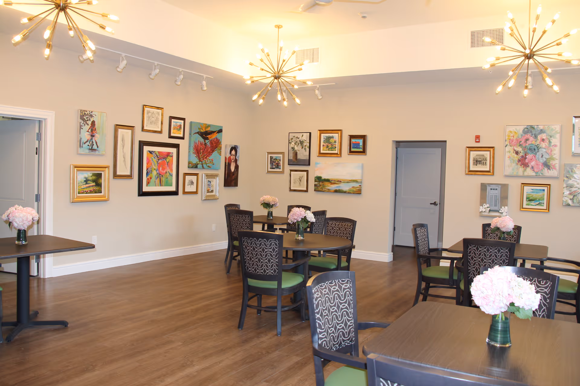 Bright communal dining room with round tables and chairs, floral centerpieces, wall art, and modern chandeliers.