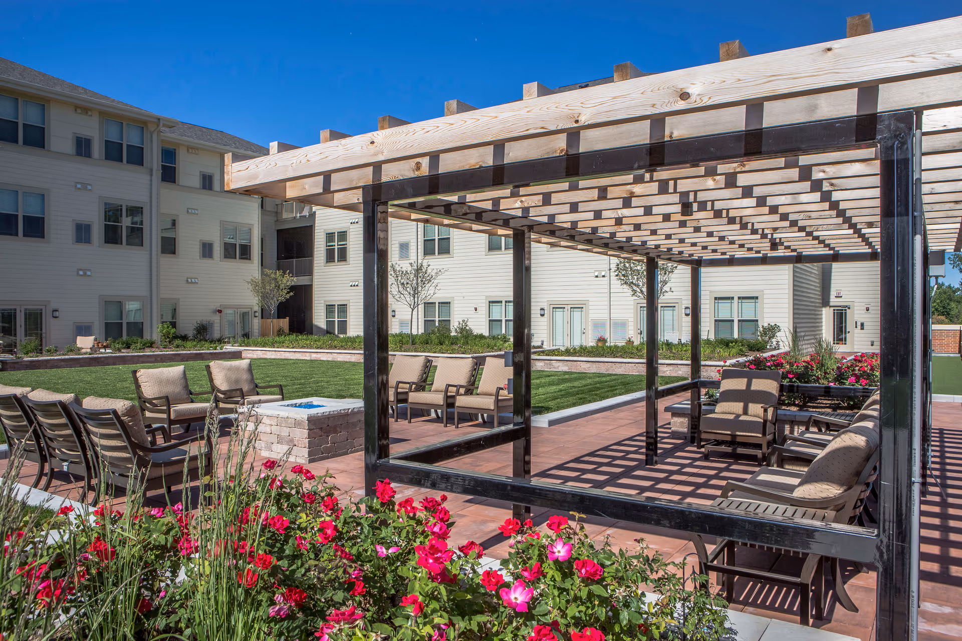 Outdoor seating area at Deer Park Village Senior Living featuring cushioned chairs arranged around a fire pit, a pergola providing partial shade, well-maintained green lawn, colorful flowers in the foreground, and a multi-story building in the background under a clear blue sky.