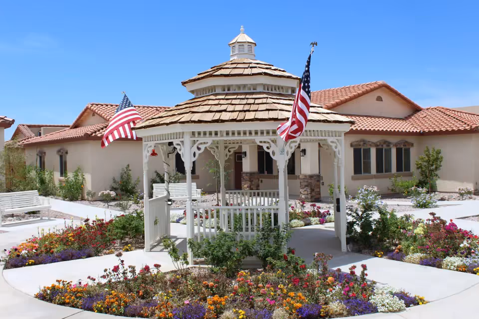 A white gazebo with a wooden shingle roof and two American flags is centered in a colorful garden with various flowers. The gazebo is surrounded by paved walkways and white benches. In the background, there is a beige building with a red tile roof under a clear blue sky.