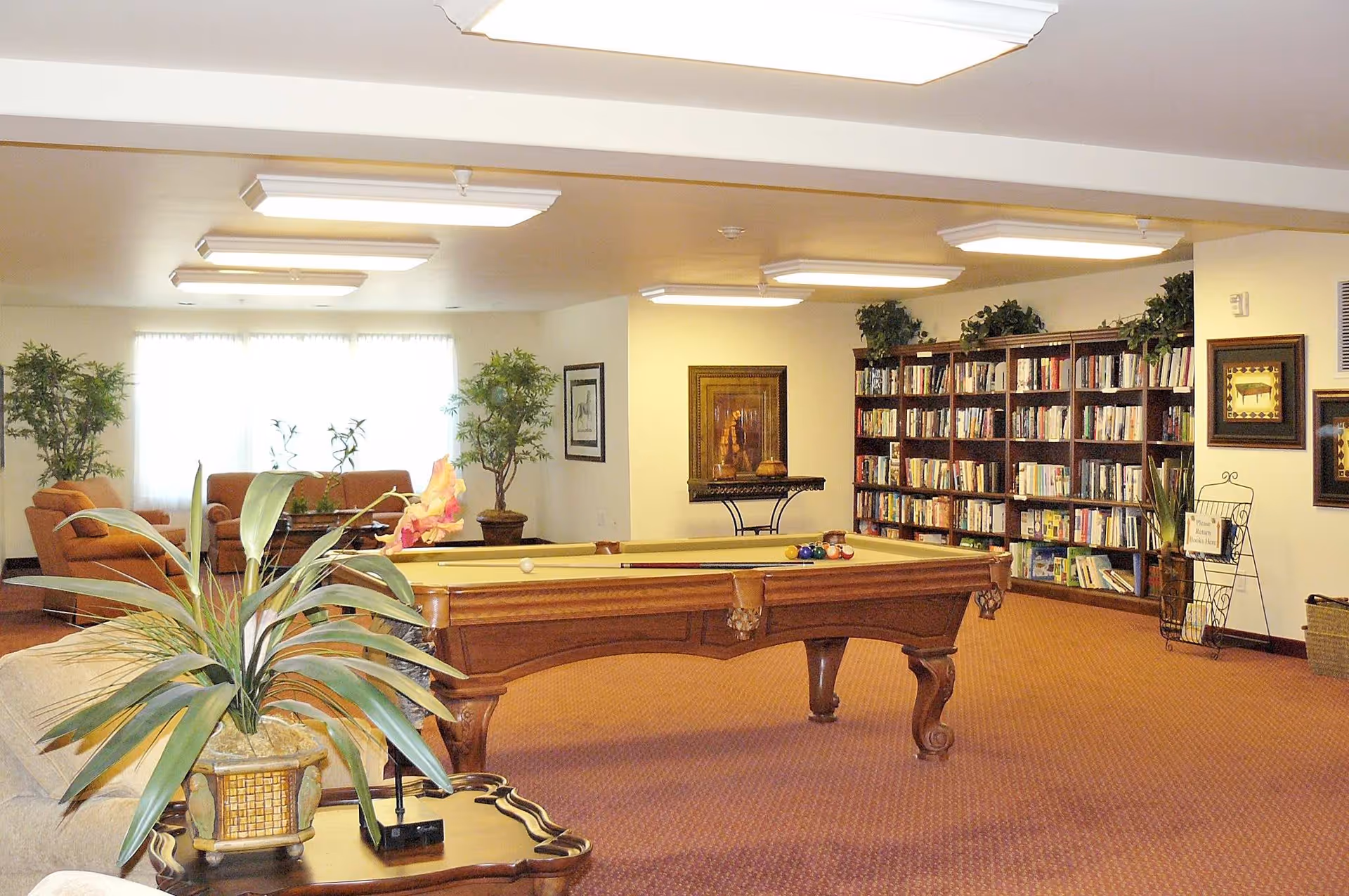 A spacious common area with a pool table in the center, surrounded by bookshelves filled with books, potted plants, framed artwork on the walls, and comfortable seating including sofas and armchairs near a large window with sheer curtains.