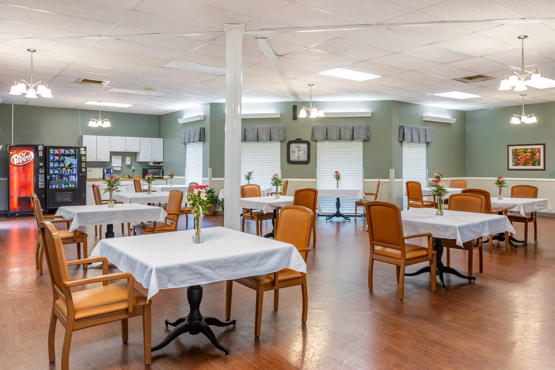A spacious dining room with multiple tables covered in white tablecloths, each adorned with a small vase of flowers. The room has wooden floors, green walls with white wainscoting, and several windows with gray valances. There are comfortable tan chairs around the tables, ceiling lights, a wall clock, a vending machine, and a snack machine in the corner near a small kitchenette area.