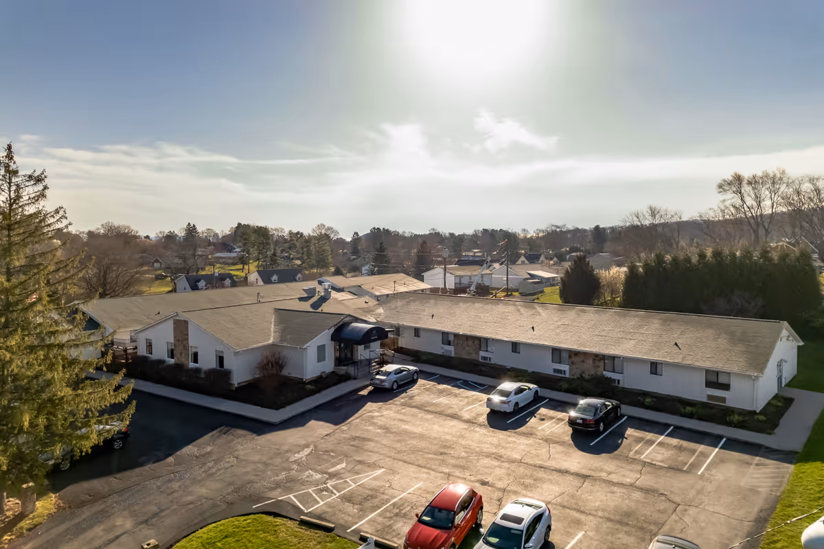 Aerial view of a single-story senior living facility building with a parking lot in front. Several cars are parked in the lot, and the building is surrounded by trees and residential houses under a partly cloudy sky with the sun shining brightly.