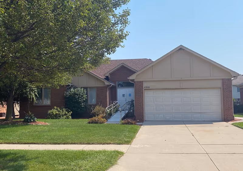 Front exterior of a single-story brick suburban house with an attached two-car garage, driveway, lawn, and a large tree.