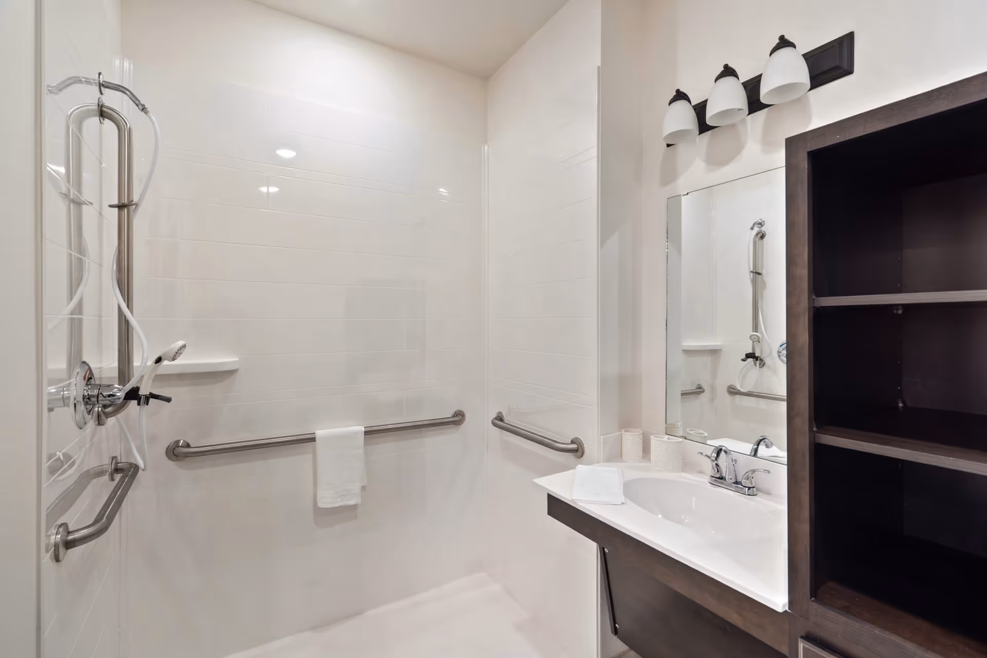 A clean and modern bathroom with white tiled walls, a walk-in shower equipped with stainless steel grab bars and a handheld showerhead. There is a white towel hanging on one of the grab bars. To the right, there is a white sink with a chrome faucet, a large mirror above it, and a dark wooden cabinet with open shelves. Above the mirror, there are three light fixtures with frosted glass shades.