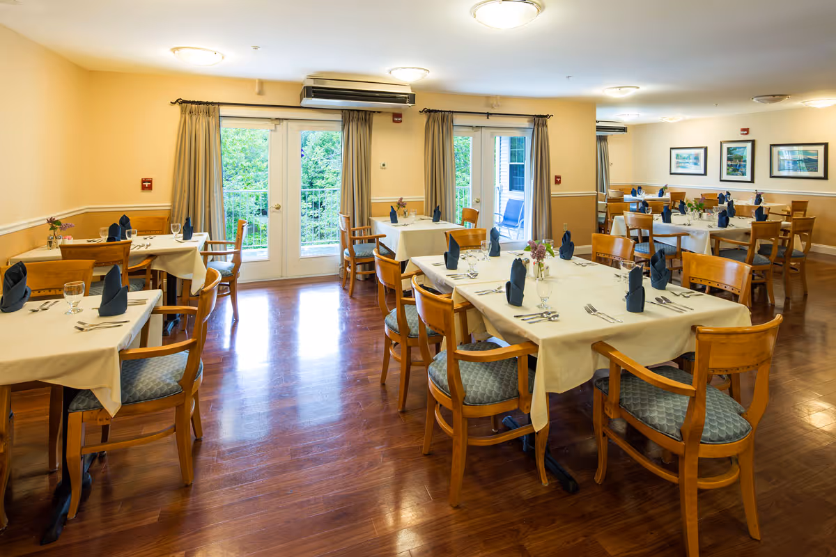 A bright dining room with several wooden tables covered with white tablecloths, each set with silverware, glasses, and folded dark blue napkins. Wooden chairs with blue cushioned seats surround the tables. Large windows and glass doors with beige curtains let in natural light and show greenery outside. The room has polished wooden floors, light yellow walls, and framed pictures on the far wall.