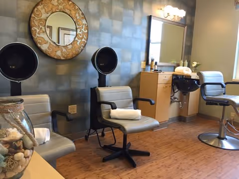 Interior of a salon area with two hair drying chairs, two salon chairs, a round decorative mirror on the wall, a sink for washing hair, and a cabinet with a mirror and light fixture above it. The floor is wooden and the walls have a textured gray finish.