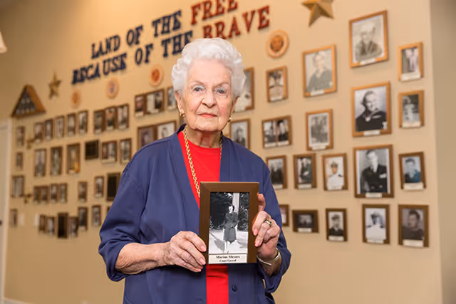 An elderly woman with white hair wearing a blue cardigan and red shirt stands indoors holding a framed black and white photograph. Behind her is a wall covered with numerous framed photos and the words 'LAND OF THE FREE BECAUSE OF THE BRAVE' displayed prominently.