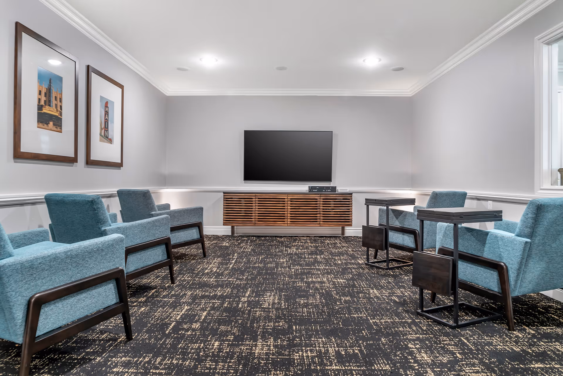 A modern lounge area with six blue upholstered armchairs arranged in two rows facing a wall-mounted flat screen TV. Below the TV is a wooden media console with horizontal slats. The room has light gray walls, two framed pictures on the left wall, recessed ceiling lights, and a patterned dark carpet.