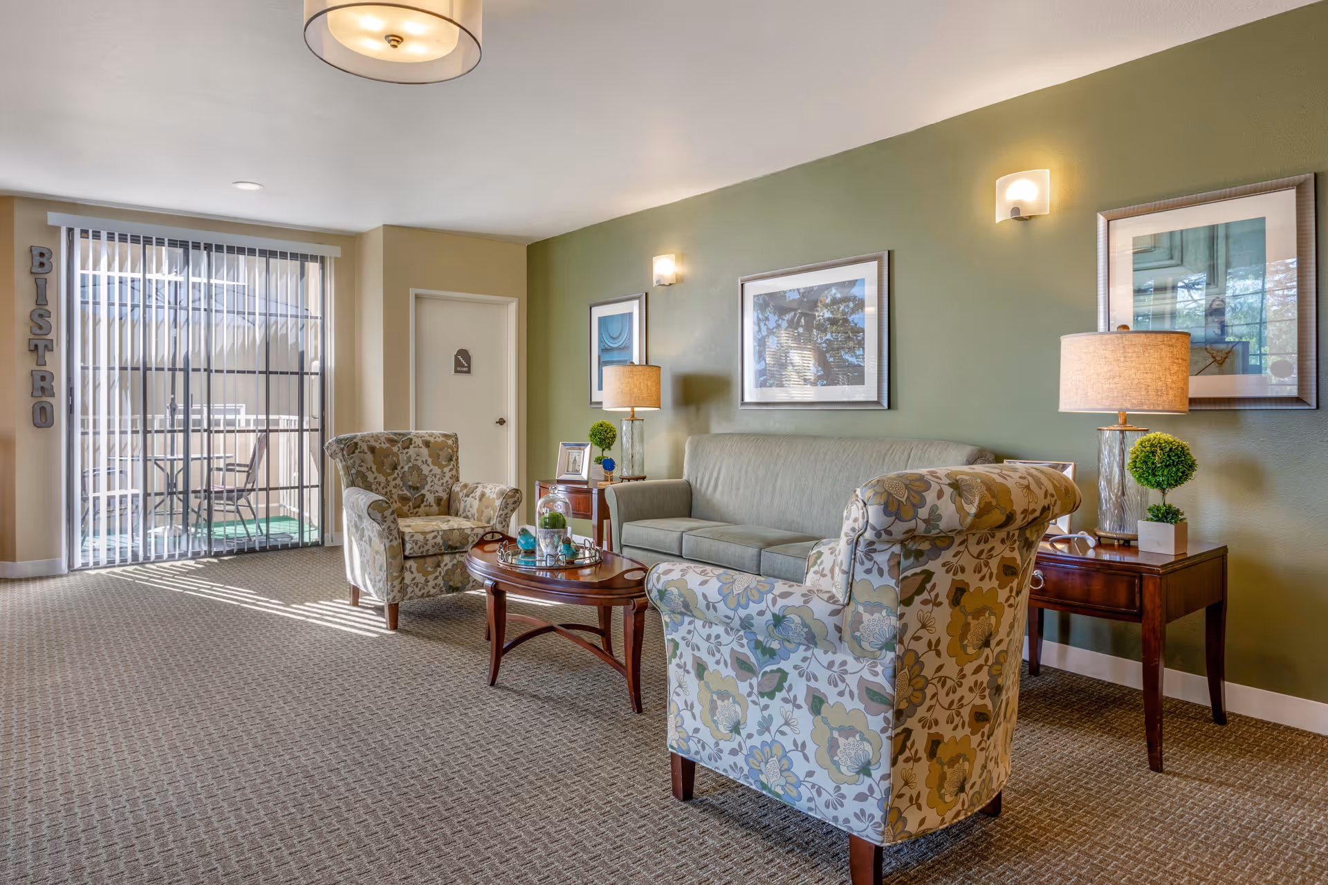 A cozy sitting area with a green sofa and two floral patterned armchairs around a wooden coffee table. The walls are painted green with framed artwork and wall sconces. There are two side tables with lamps and small decorative plants. A sliding glass door with vertical blinds leads to an outdoor patio with a table and chairs. The word 'BISTRO' is displayed vertically on the wall near the door.