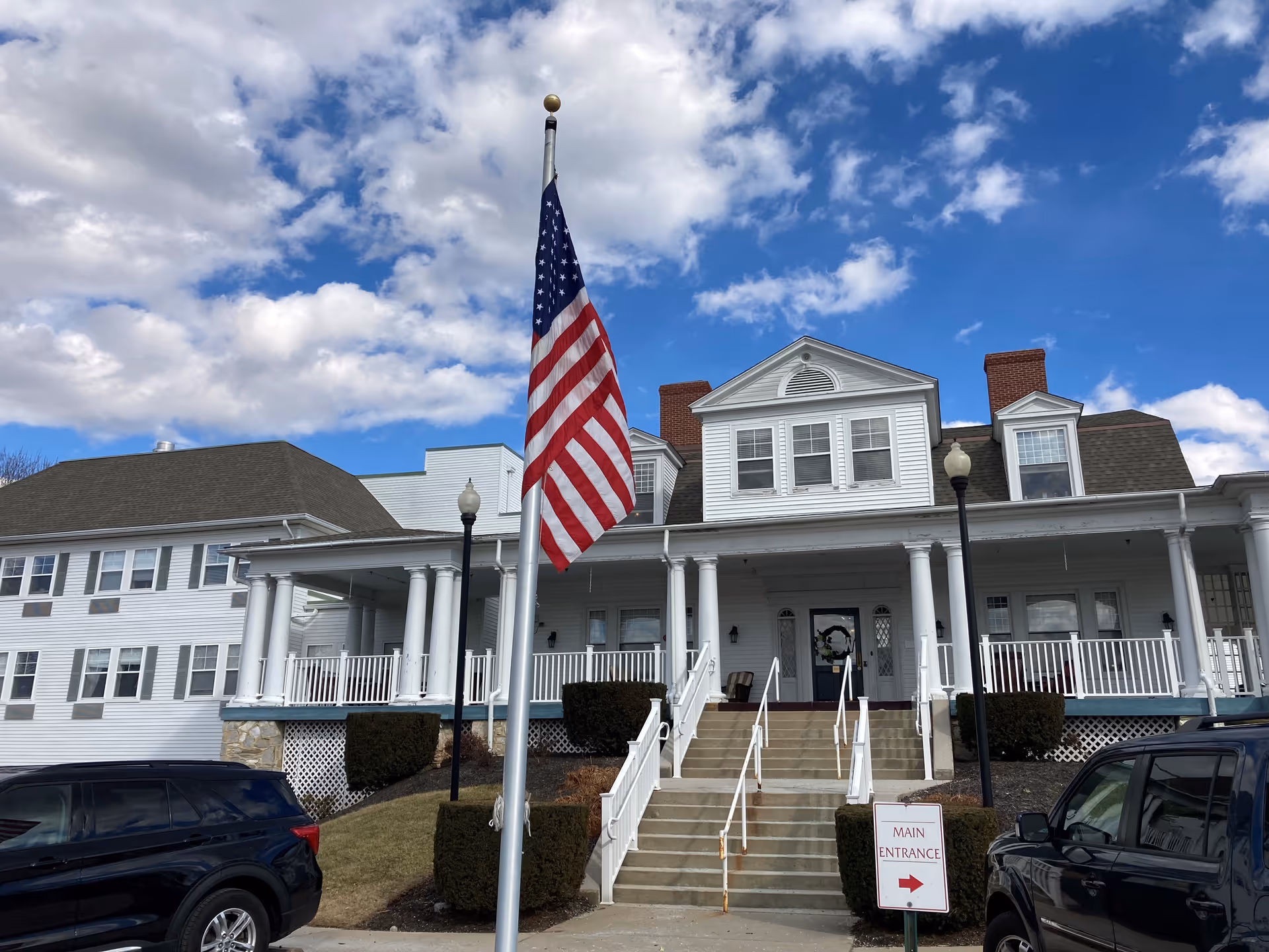 Front exterior view of a large white senior living facility building with a porch, stairs leading to the main entrance, two black lampposts, an American flag on a flagpole, and two parked cars on either side of the entrance. The sky is partly cloudy with blue patches.