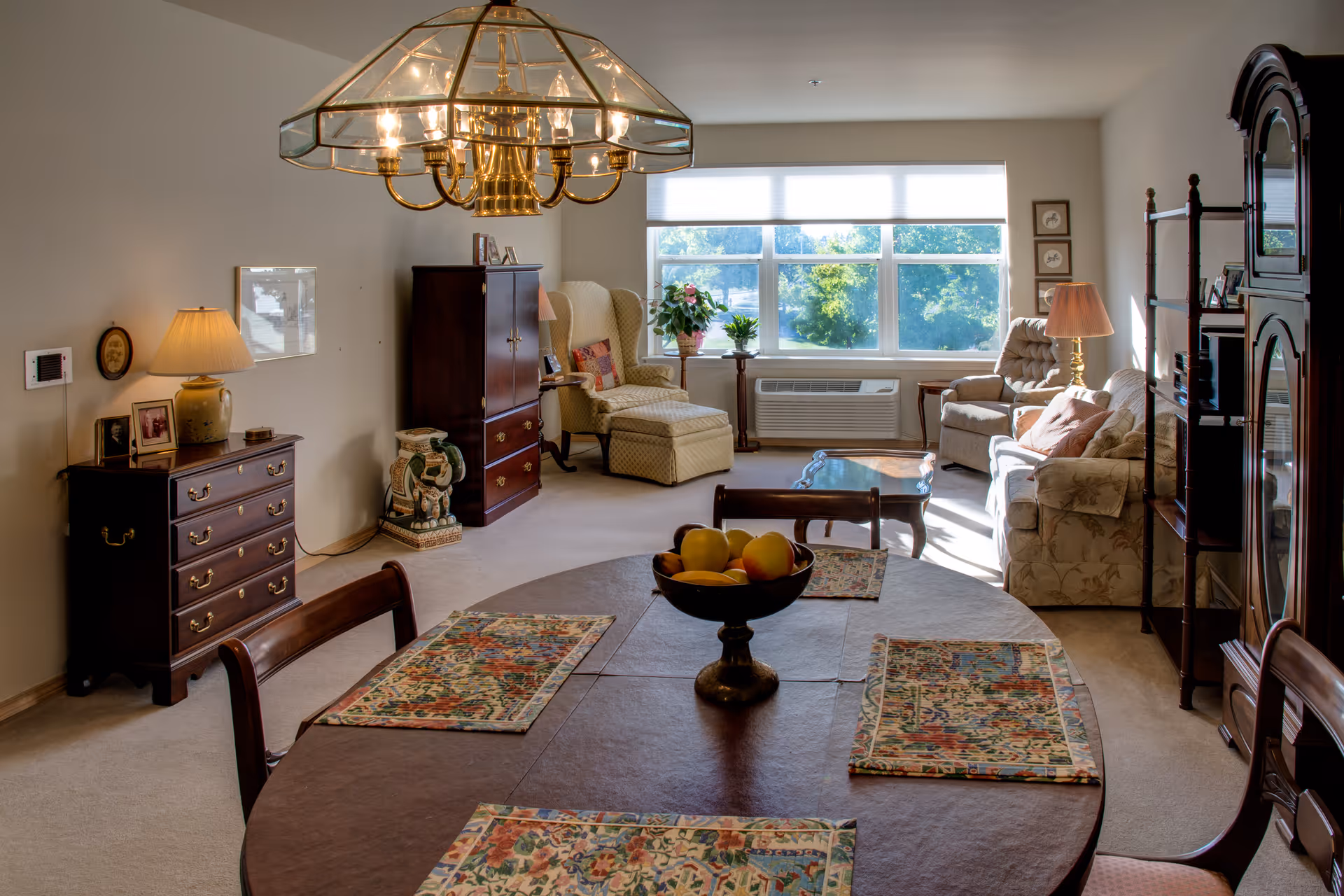 Sunlit combined living and dining room with a round table set with placemats and a fruit bowl, upholstered chairs, wooden cabinets, and a large window.