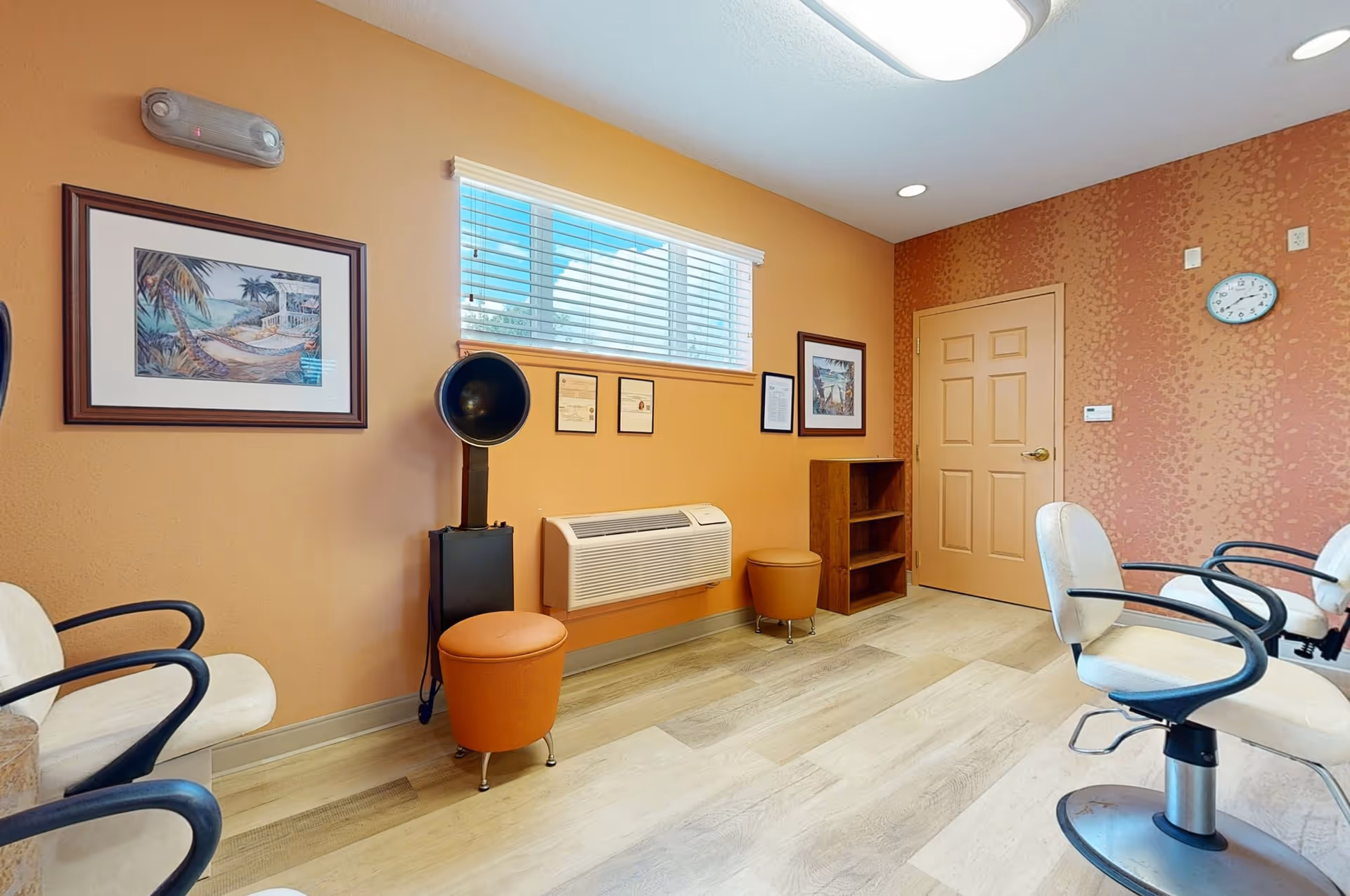Interior of a salon or beauty room with orange walls, a window with blinds, framed pictures on the walls, salon chairs with armrests, a hair dryer hood, two small round stools, a wooden shelf, and a wall-mounted air conditioning unit.