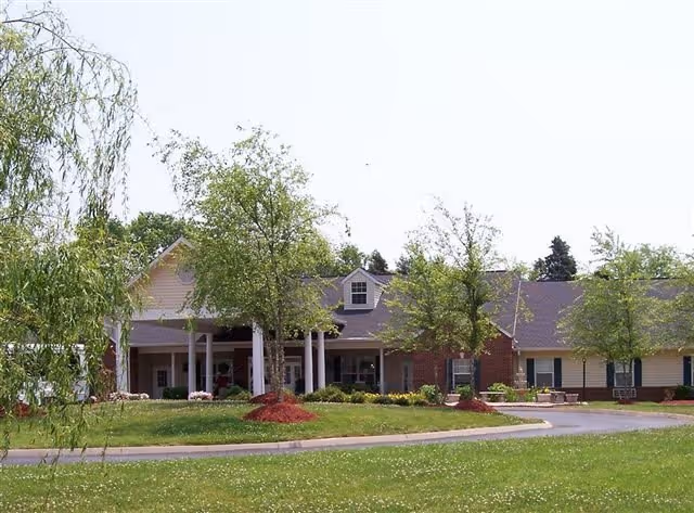 Exterior view of a single-story senior living facility building with a covered entrance, surrounded by green grass, trees, and landscaping under a clear sky.