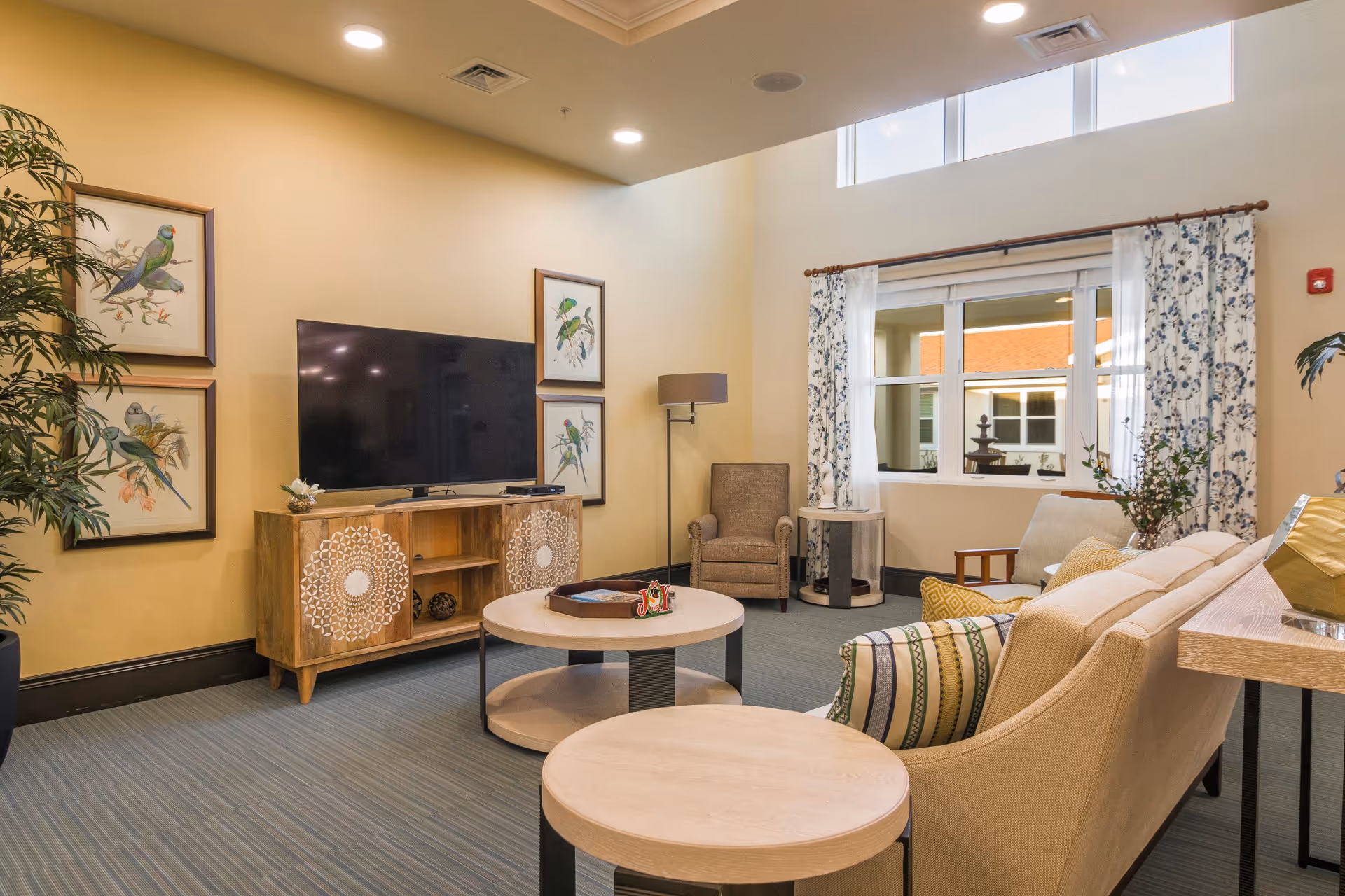 A cozy living room with a beige sofa adorned with patterned pillows, a round coffee table, and a matching side table. A wooden TV stand with intricate designs holds a large flat-screen TV. The walls are decorated with framed bird artwork, and a tall floor lamp stands near a single armchair. Large windows with floral curtains allow natural light to brighten the room.