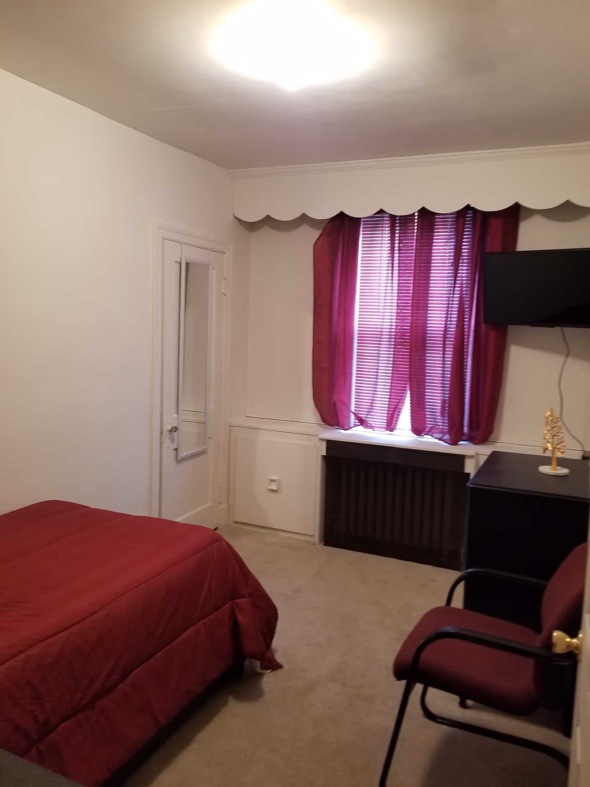 A small bedroom with a bed covered in a red comforter, a window with red curtains and blinds, a wall-mounted TV, a black dresser with a decorative item on top, and a maroon chair with black armrests. The walls are white and there is a door with a mirror on it.