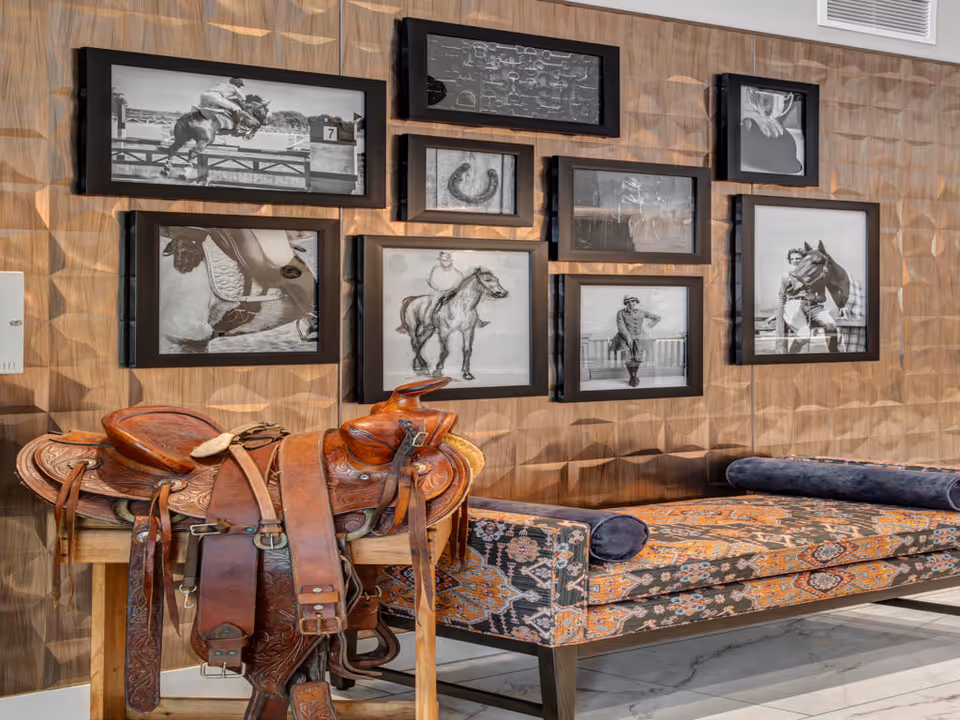 Interior room with a patterned upholstered bench and a wooden saddle stand holding a detailed leather saddle. The wall behind features a textured geometric design and is decorated with multiple black and white framed photographs and artwork, mostly depicting horses and equestrian themes.