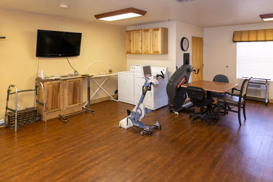 A multipurpose room with wooden flooring featuring a wall-mounted TV above a wooden cabinet, an ironing board, a walker, a stationary exercise bike, a rowing machine, a table with four chairs, a clock on the wall, a window with blinds and a valance, and a washing machine and dryer under wooden cabinets.
