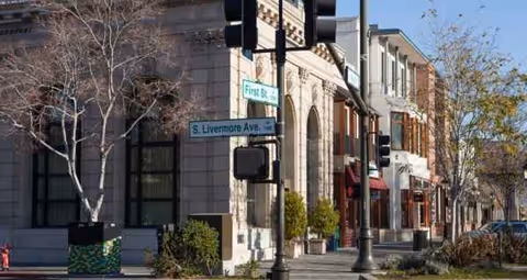 Exterior street view with brick storefronts, trees, and traffic lights along a sunlit sidewalk.