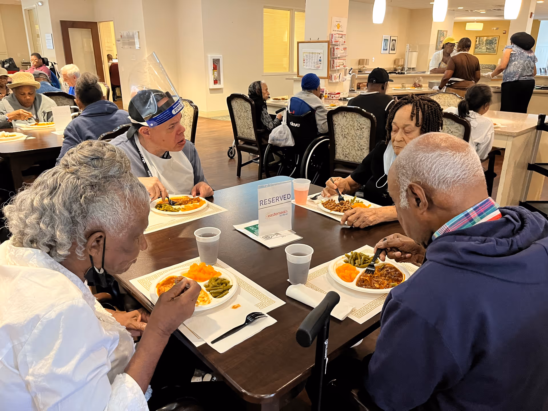 A group of older adults seated around a dining table in a communal dining room eating meals.