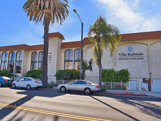 Exterior view of Villa Redondo Assisted Living Home building with large windows, palm trees, and parked cars along the street in front.