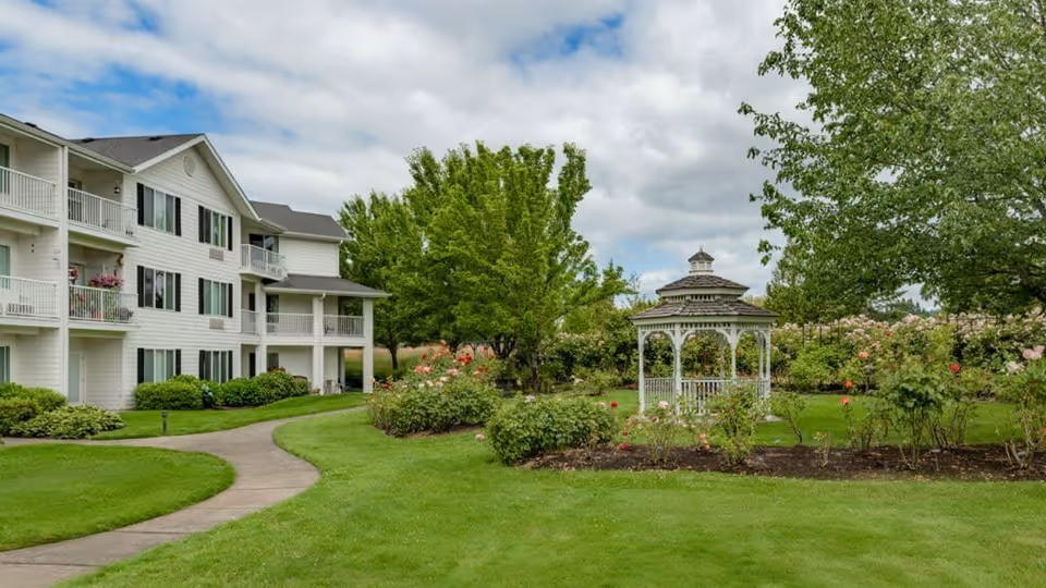 A well-maintained garden area with a white gazebo surrounded by rose bushes and green trees. A paved walkway curves through the green lawn leading to a three-story white residential building with balconies and windows. The sky is partly cloudy.