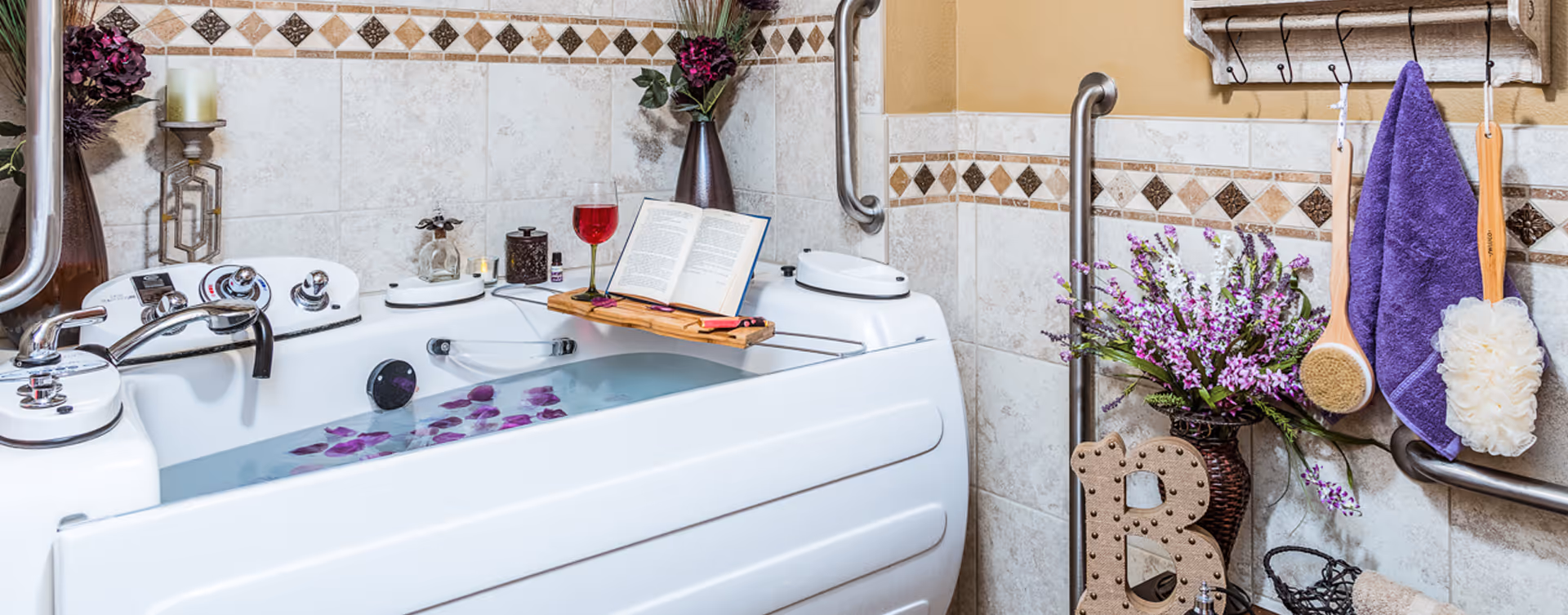 A white bathtub filled with water and purple flower petals in a tiled bathroom. The tub has metal grab bars on the walls for support. A wooden bath tray holds an open book and a glass of red wine. There are decorative items including a vase with purple flowers, a candle, and bath brushes hanging on the wall. A purple towel is also hanging on hooks above the tub.
