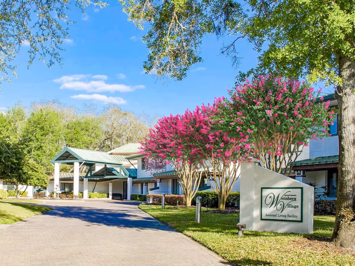 Driveway and entrance of Weinberg Village Assisted Living with a sign, green-roofed portico, and pink flowering trees.