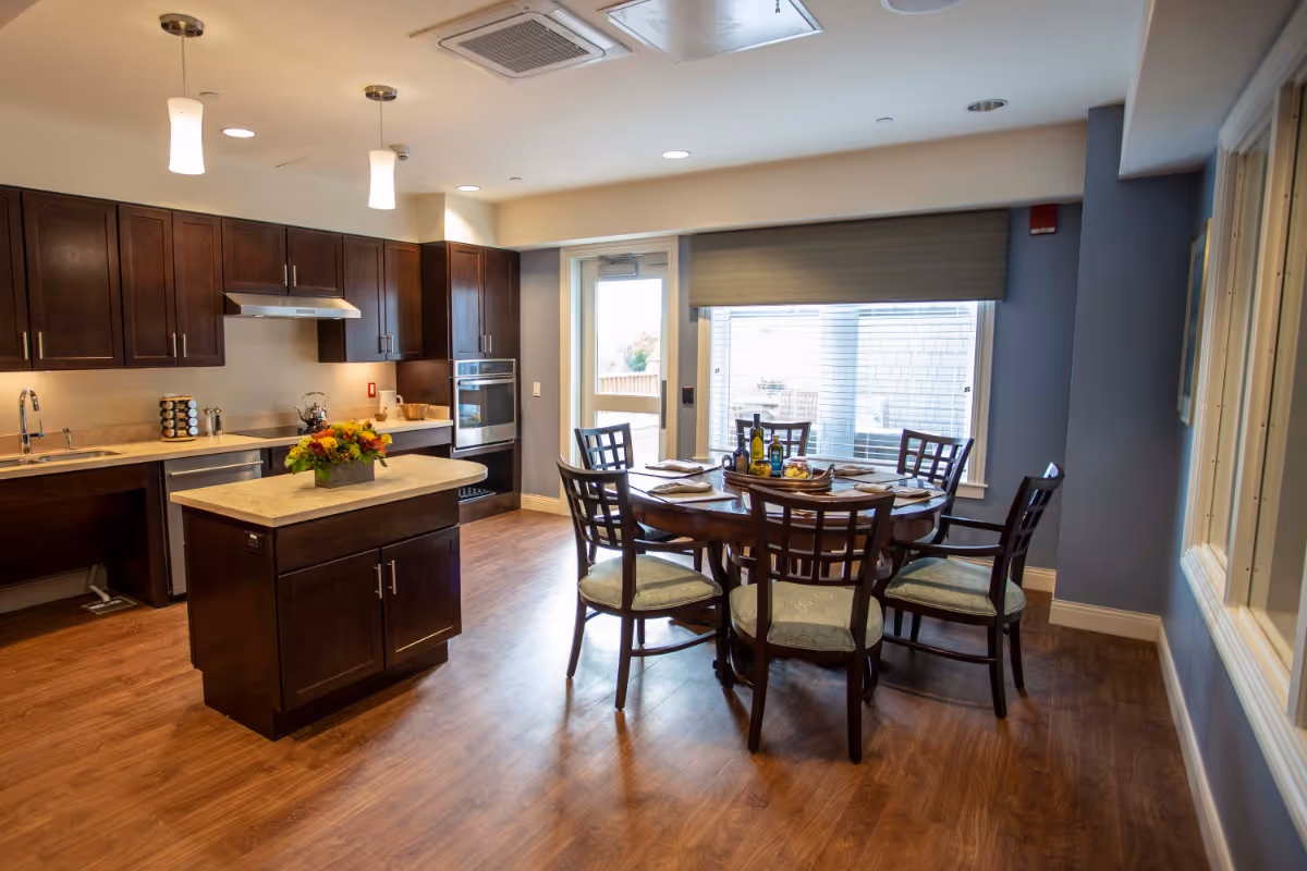 A bright and modern kitchen and dining area with dark wood cabinets, a small kitchen island with a flower arrangement, and a round dining table set with six chairs. The room has wood flooring, pendant lights, a large window with blinds, and a glass door leading outside.