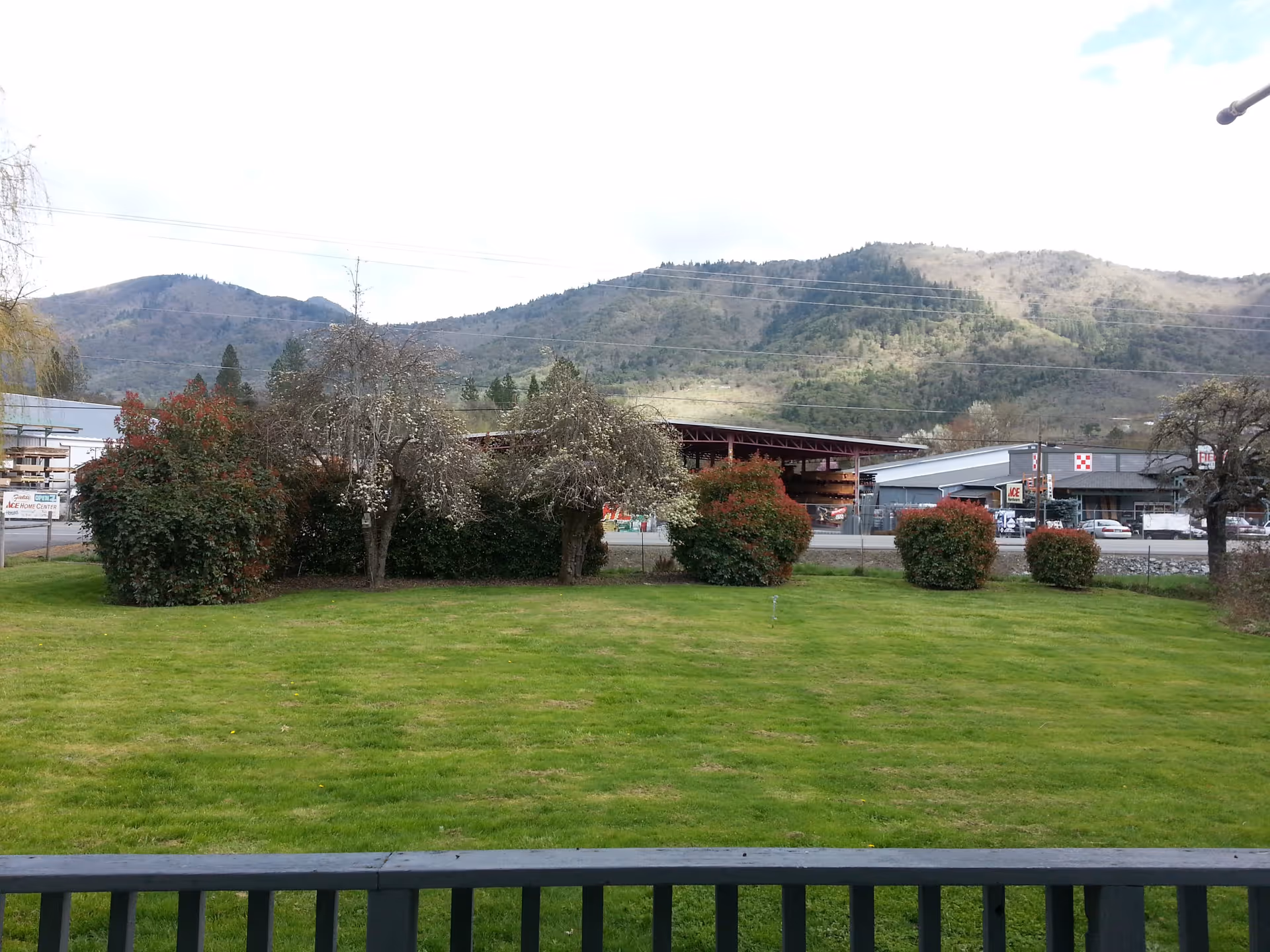 Grassy lawn and shrubs with a road and commercial buildings in front of tree-covered hills and mountains in the background.