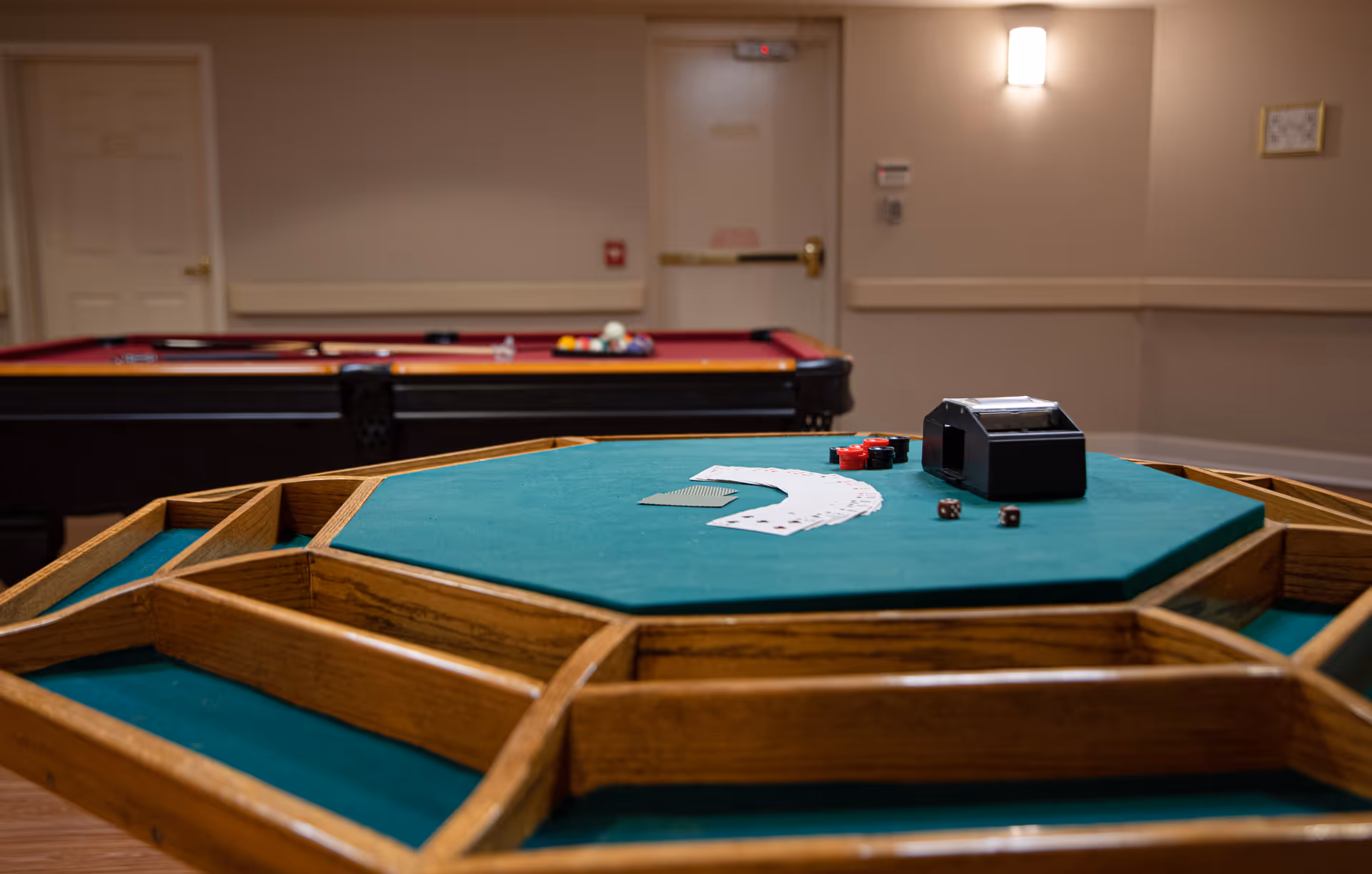 A game room featuring a green felt octagonal card table with playing cards, poker chips, dice, and a card shuffler on top. In the background, there is a pool table with balls arranged for a game. The room has beige walls, a closed door, and a wall-mounted light fixture.
