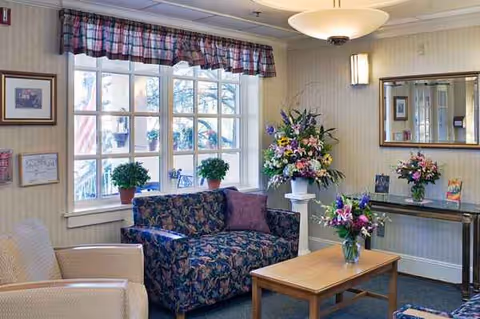Cozy senior living lounge with floral furniture, plants, and a large window.