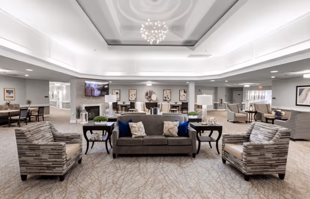 A spacious and well-lit senior living facility common area with a central seating arrangement including a gray sofa with blue and beige pillows, two patterned armchairs, and side tables with lamps and plants. The room features a high ceiling with a modern chandelier, a wall-mounted TV above a fireplace, and additional seating and tables in the background. The decor is neutral with light-colored walls and carpet.