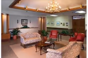 A well-lit living room area with two floral-patterned sofas, two red armchairs, a wooden coffee table, and potted plants. The room features a chandelier, framed artwork on the walls, and a reception desk in the background.