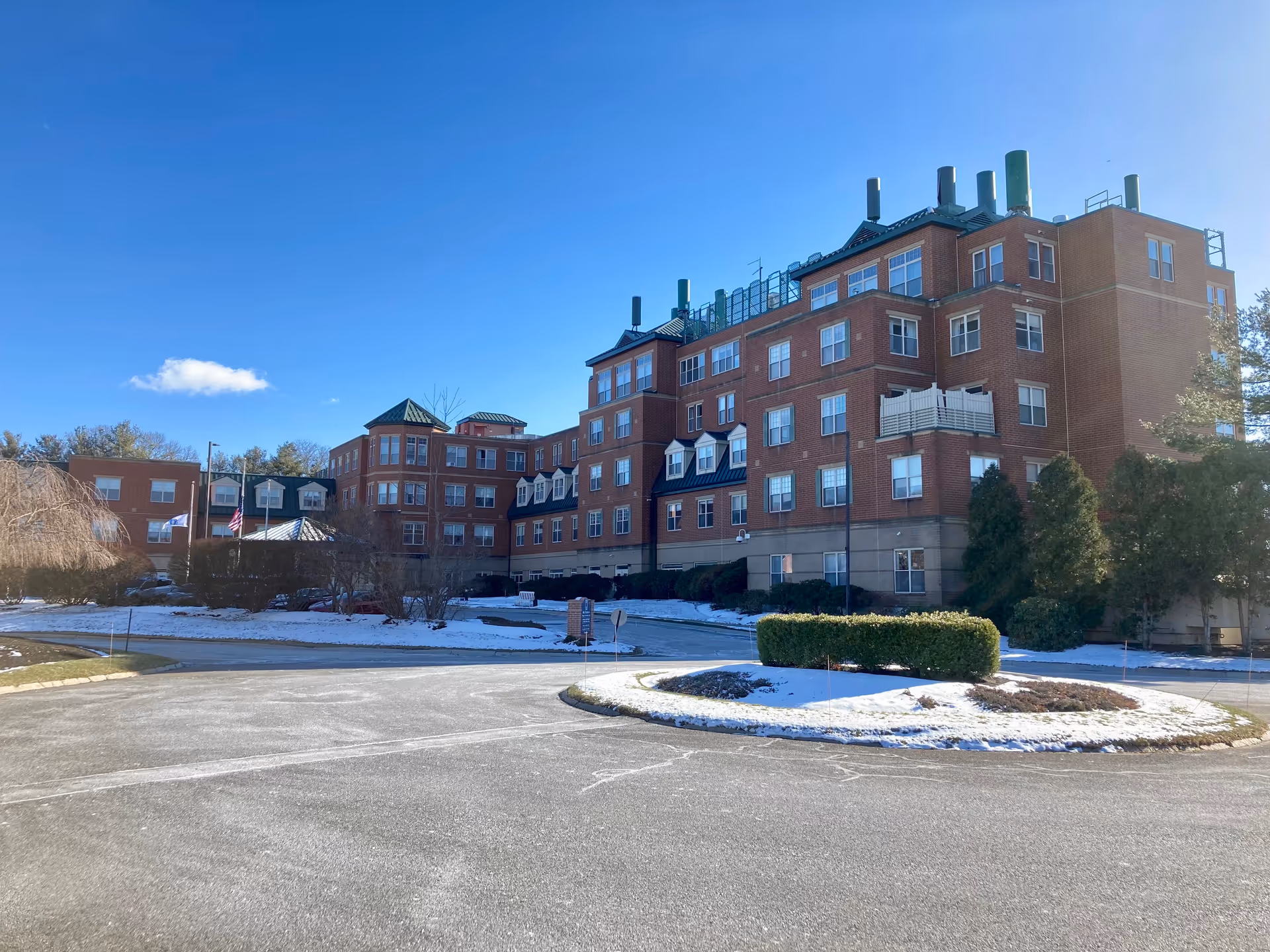Large multi-story red-brick senior living building with snow-dusted landscaping under a clear blue sky.