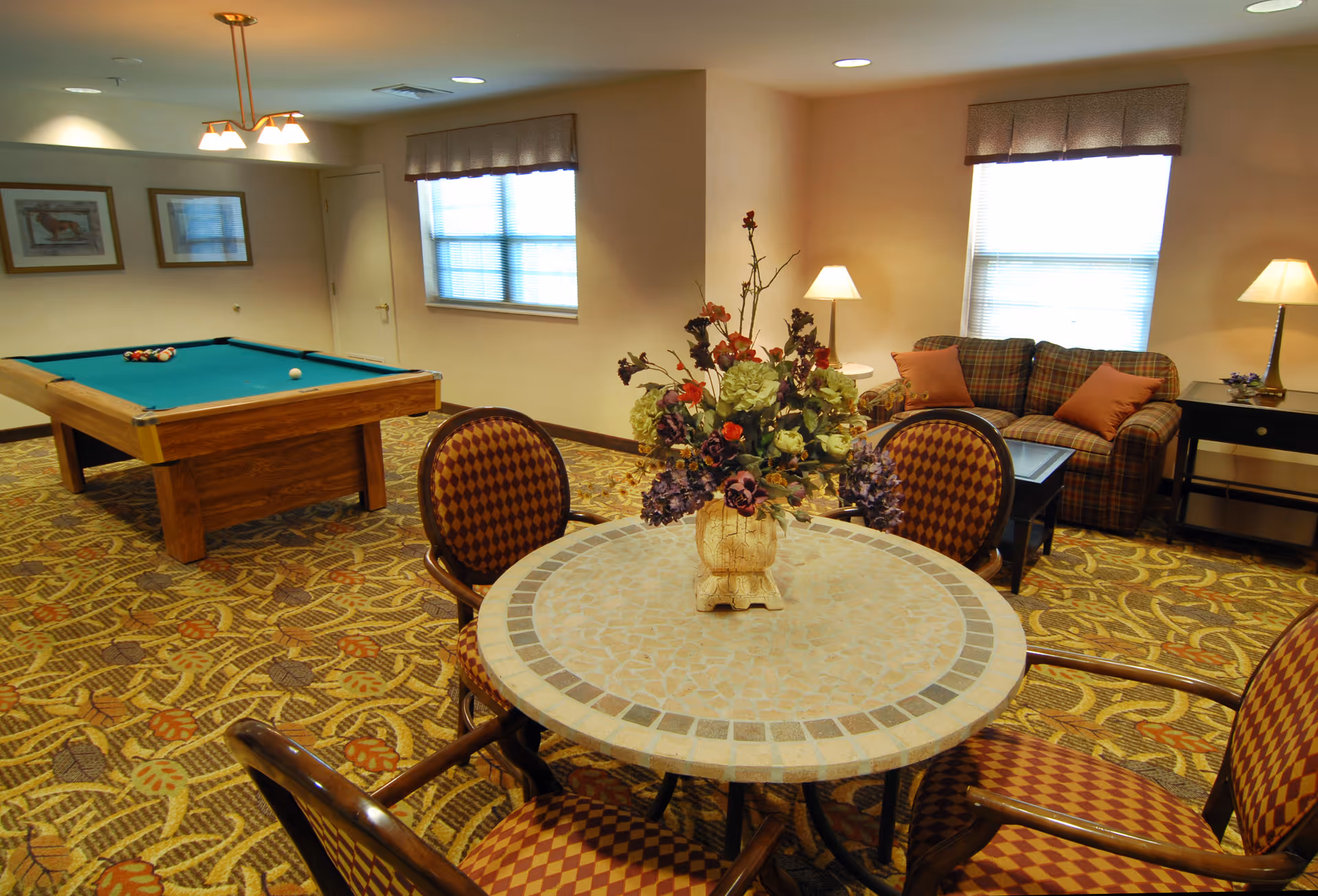 A cozy common room featuring a round tiled table with a floral centerpiece, patterned chairs, a pool table, and a sofa seating area.