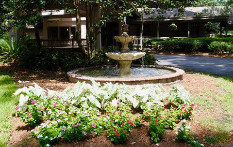 Outdoor garden area with a tiered water fountain surrounded by a circular stone border. In front of the fountain, there are various flowering plants and greenery. Trees and a building with a covered walkway are visible in the background.