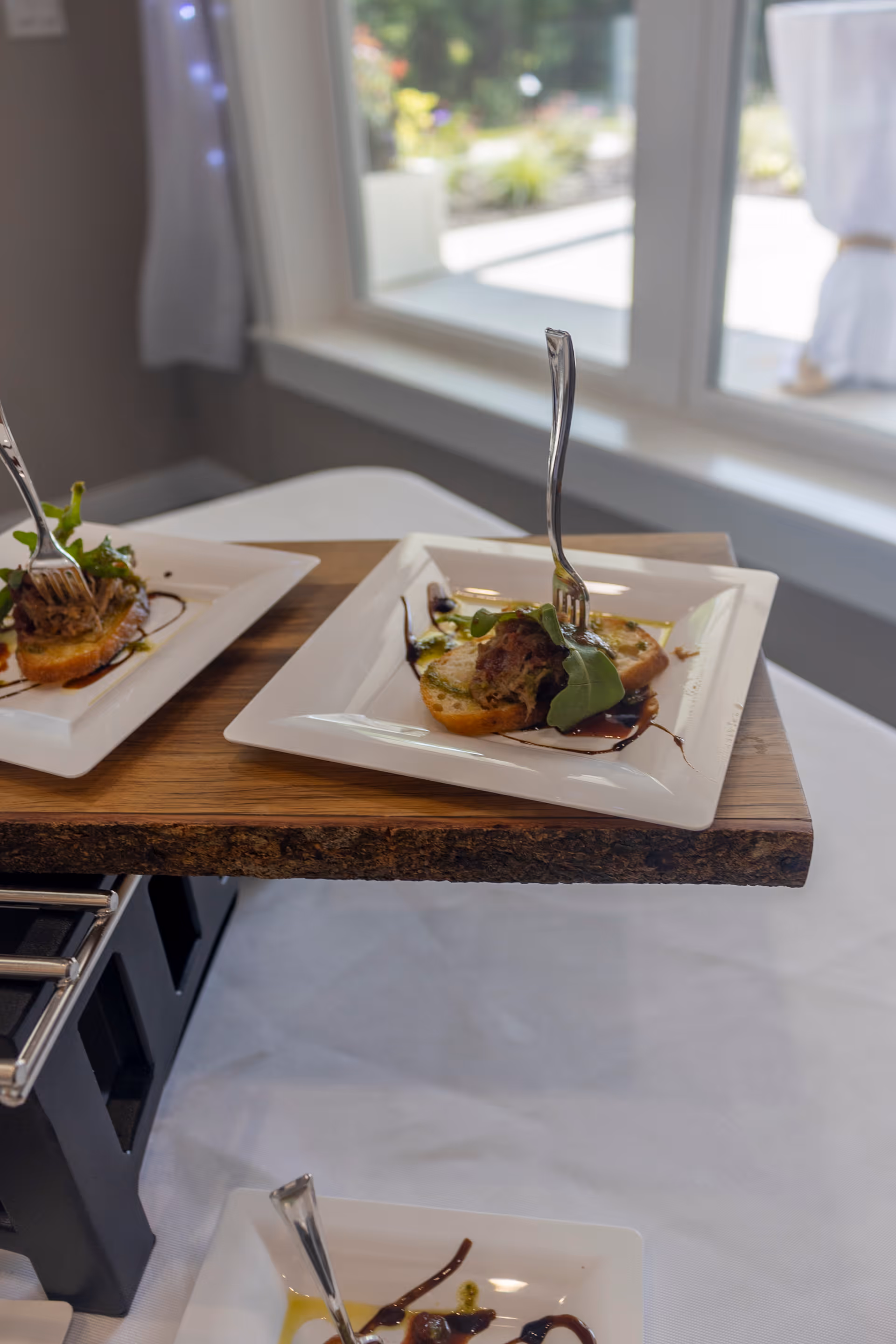 Close-up of three white square plates with gourmet appetizers, each featuring a small portion of food garnished with greens and drizzled with sauce, placed on a wooden serving board near a window with natural light.