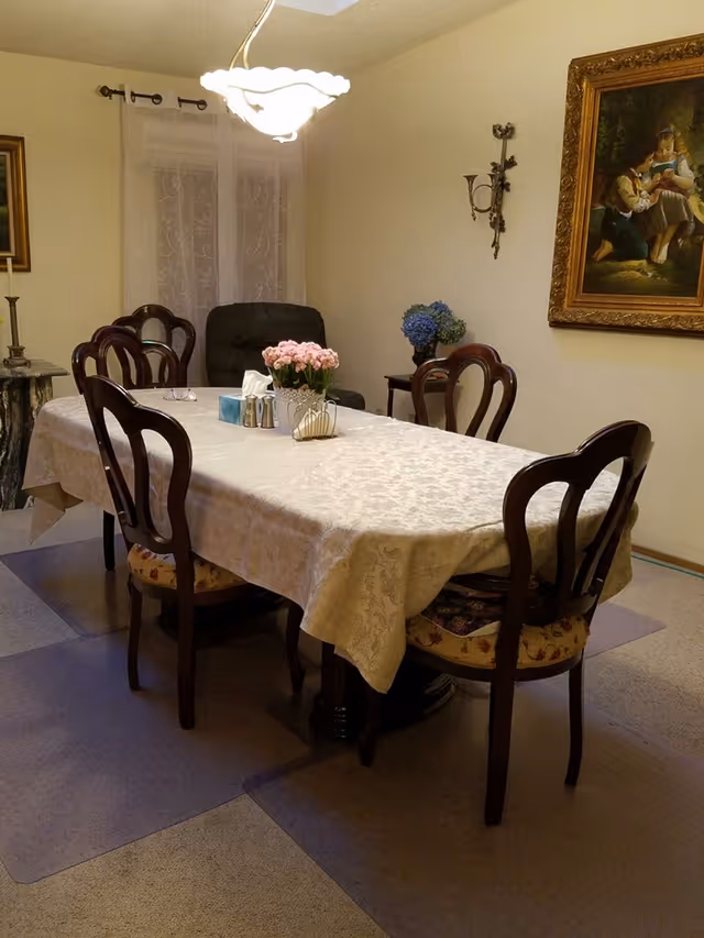 A dining room with a rectangular table covered with a light-colored tablecloth. Six wooden chairs with patterned cushions surround the table. On the table, there is a flower pot with pink flowers, a tissue box, salt and pepper shakers, and a napkin holder. The room has a hanging light fixture above the table, a framed painting on the wall, and a small side table with a blue flower arrangement.