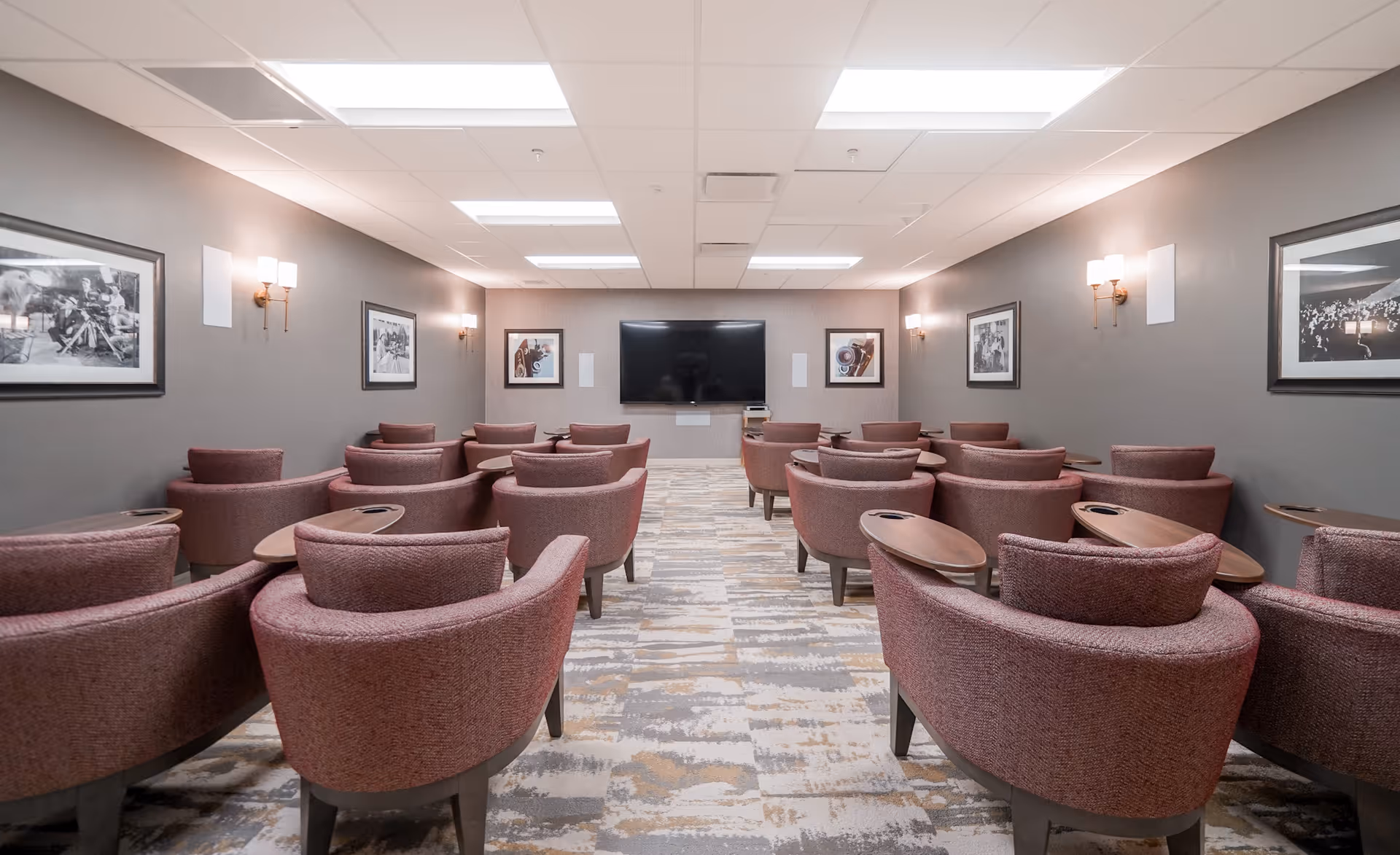 A room with multiple rows of upholstered chairs, each with a small attached wooden table, facing a large flat-screen TV mounted on the far wall. The walls are painted gray and decorated with framed black and white photographs. The ceiling has recessed lighting panels and wall sconces provide additional light.