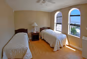 A simple bedroom with two single beds covered in white bedspreads, a wooden nightstand with a white lamp between the beds, light-colored walls, wooden flooring, and two arched windows letting in natural light.