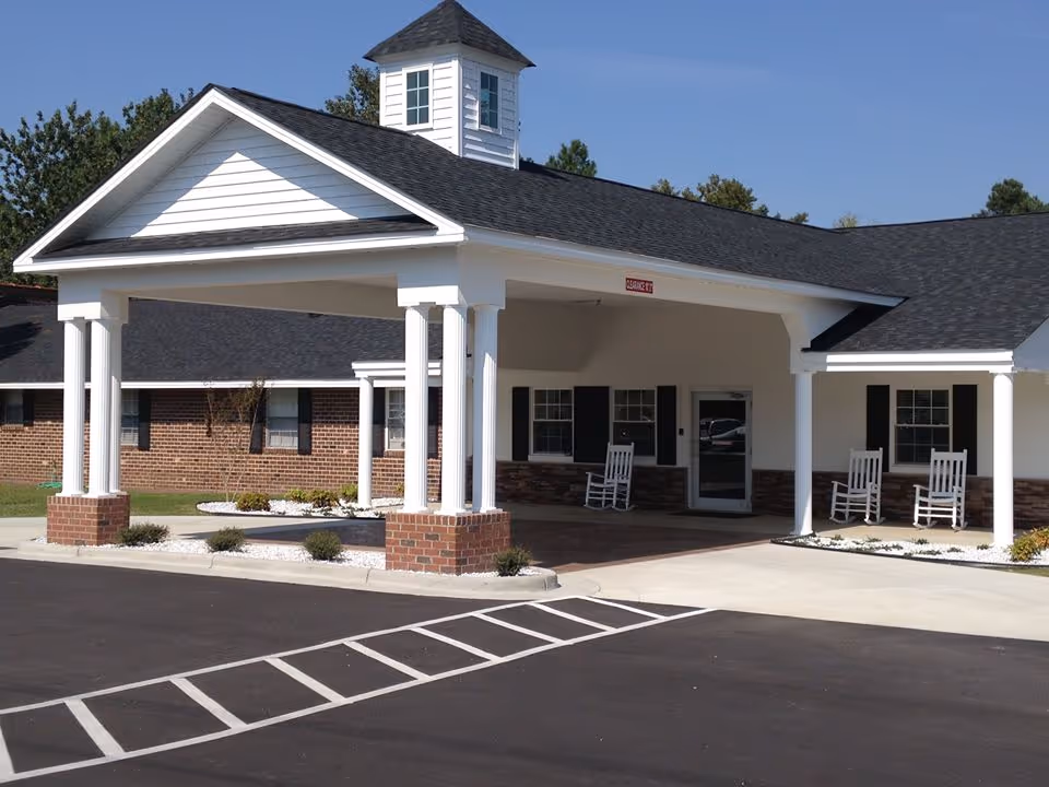 Entrance of a single-story brick and white siding building with a covered driveway supported by white columns with brick bases. There are three white rocking chairs on the porch area near the entrance door. The sky is clear and blue.