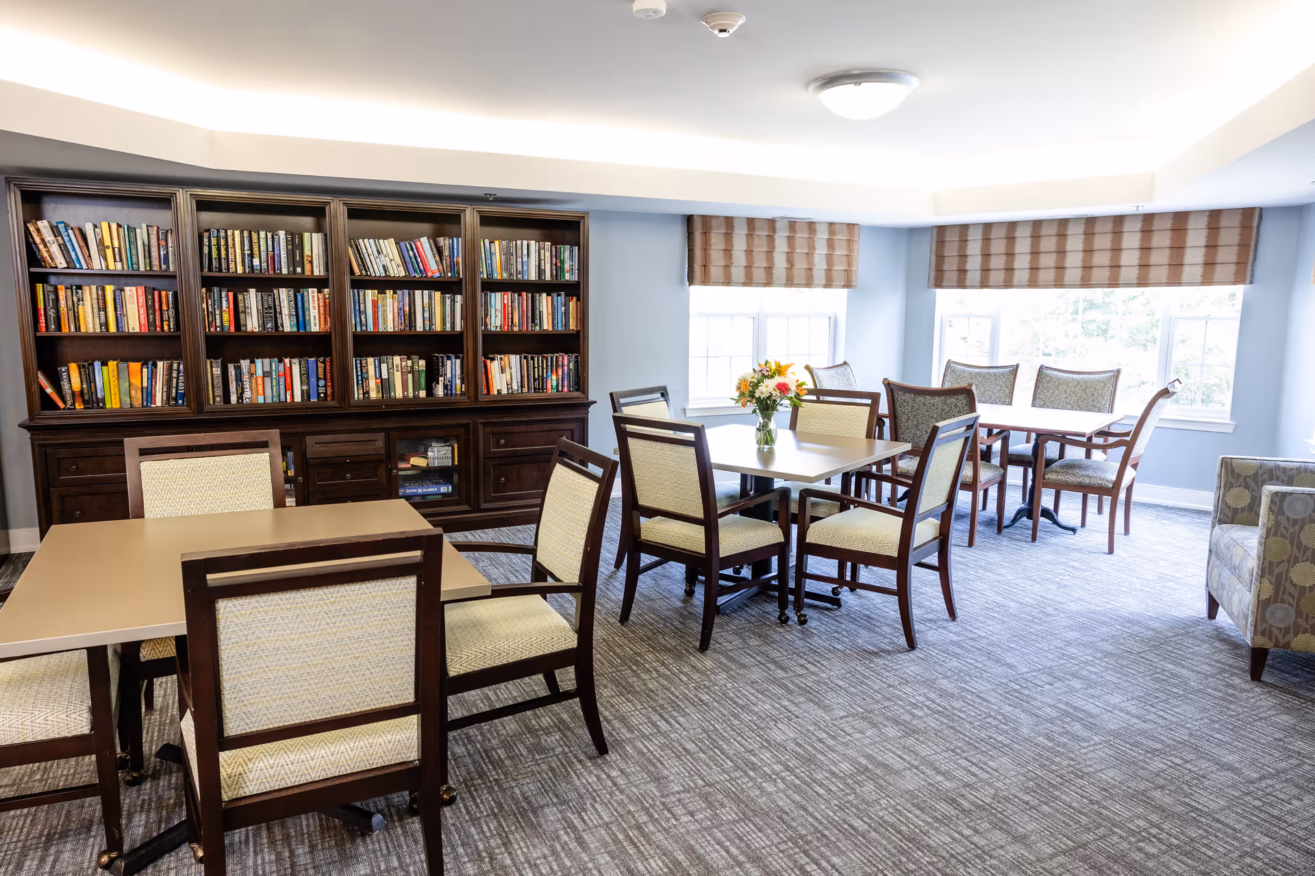 A bright room with several tables and chairs arranged for seating. There is a large wooden bookshelf filled with books against one wall. The room has large windows with striped blinds, and a vase with flowers is placed on one of the tables. The carpet is patterned in neutral tones, and there is an upholstered armchair visible on the right side.