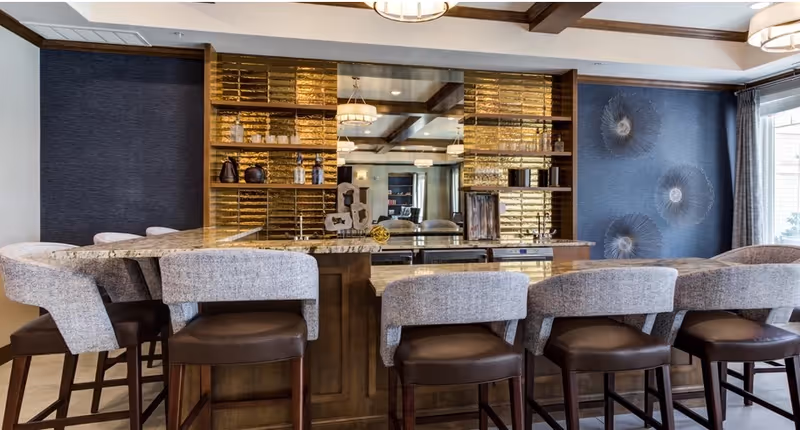 A modern senior living facility bar area with a granite countertop and six cushioned bar stools with wooden legs. Behind the bar is a mirrored wall with golden decorative tiles and shelves holding various decorative items. The room features dark blue textured walls with artistic wall hangings and large windows with curtains allowing natural light to enter.